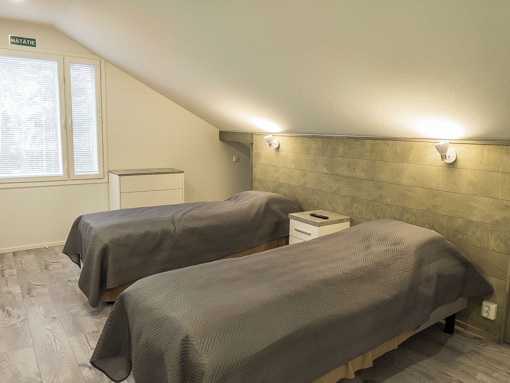 Pieni hotellihuone, jossa on yksi huone, kaksi sänkyä, yöpöytä, seinään kiinnitetty lamppu ja ikkuna, jossa on sälekaihtimet.