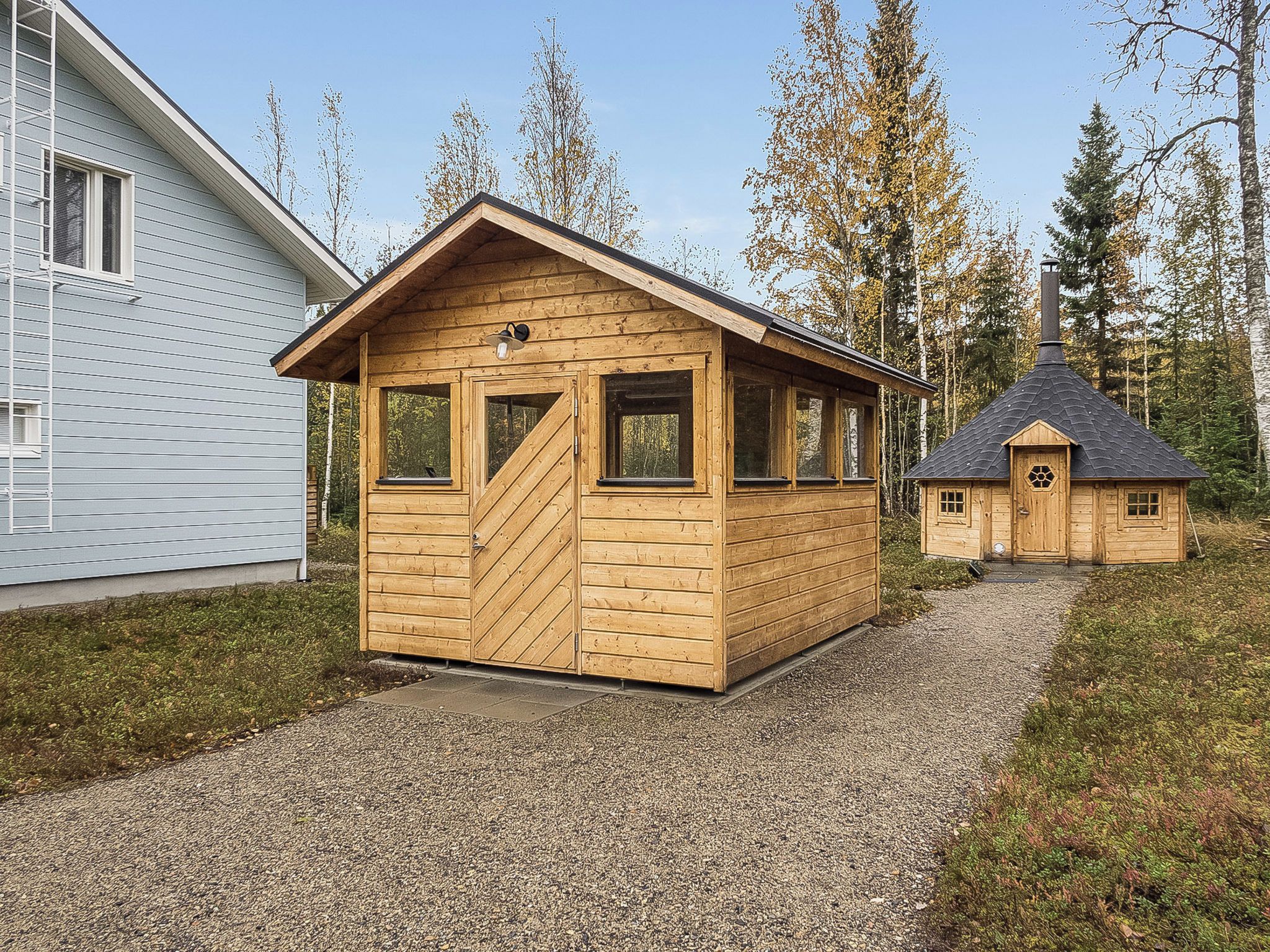 Puisesta hirsirakenteisesta mökistä, jossa on harjakatto, ovi ja ikkunat, on näkymä suurempaan taloon ja pienempään rakennukseen, jossa on savupiippu.