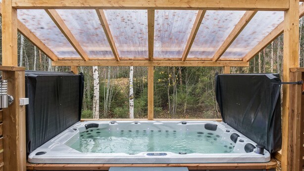 A hot tub with a wooden deck, surrounded by a wooden pergola with a mesh roof, set in a forested area.
