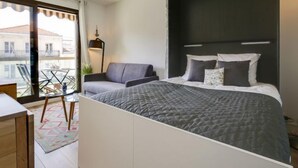 A modern bedroom with a large bed, a sofa, and a balcony with a view of buildings. - Sun Beach by Interhome (Arcachon)