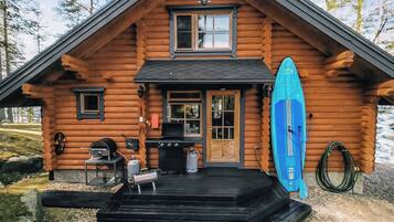 Une cabane en rondins avec une véranda, un barbecue et une planche de surf.