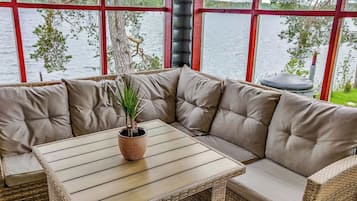 Un coin salon avec une table en osier et des bancs rembourrés, une plante en pot et une vue sur un lac, visible à travers de grandes fenêtres.