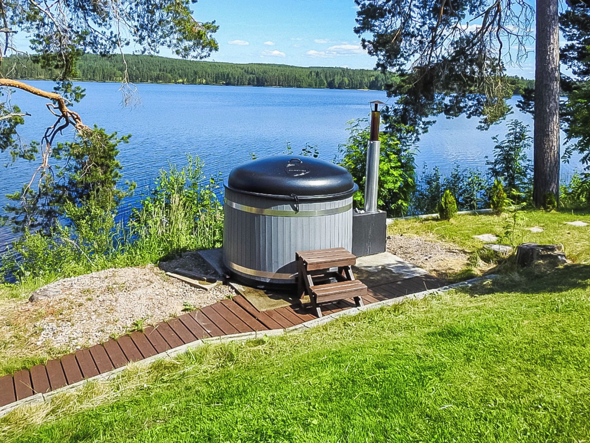 A portable grill with a chimney, a wooden bench, and a paved path leading to a lake.