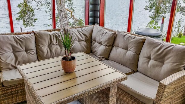 En hjørnesofa med et vævet bord foran, en potteplante og udsigt til en sø gennem store vinduer.