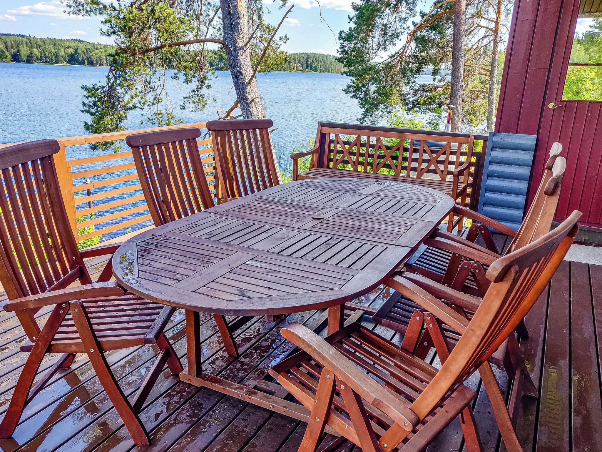 Ulkotilaan sijoitettu puinen terassi, jossa on pyöreä pöytä ja tuolit. Terassilta on näkymä vesistöön ja puustoon.