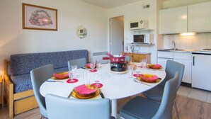 Comedor con una mesa redonda dispuesta para cuatro personas, con platos, vasos y cubiertos. Se ve un sofá azul y un microondas en la zona de la cocina al fondo.