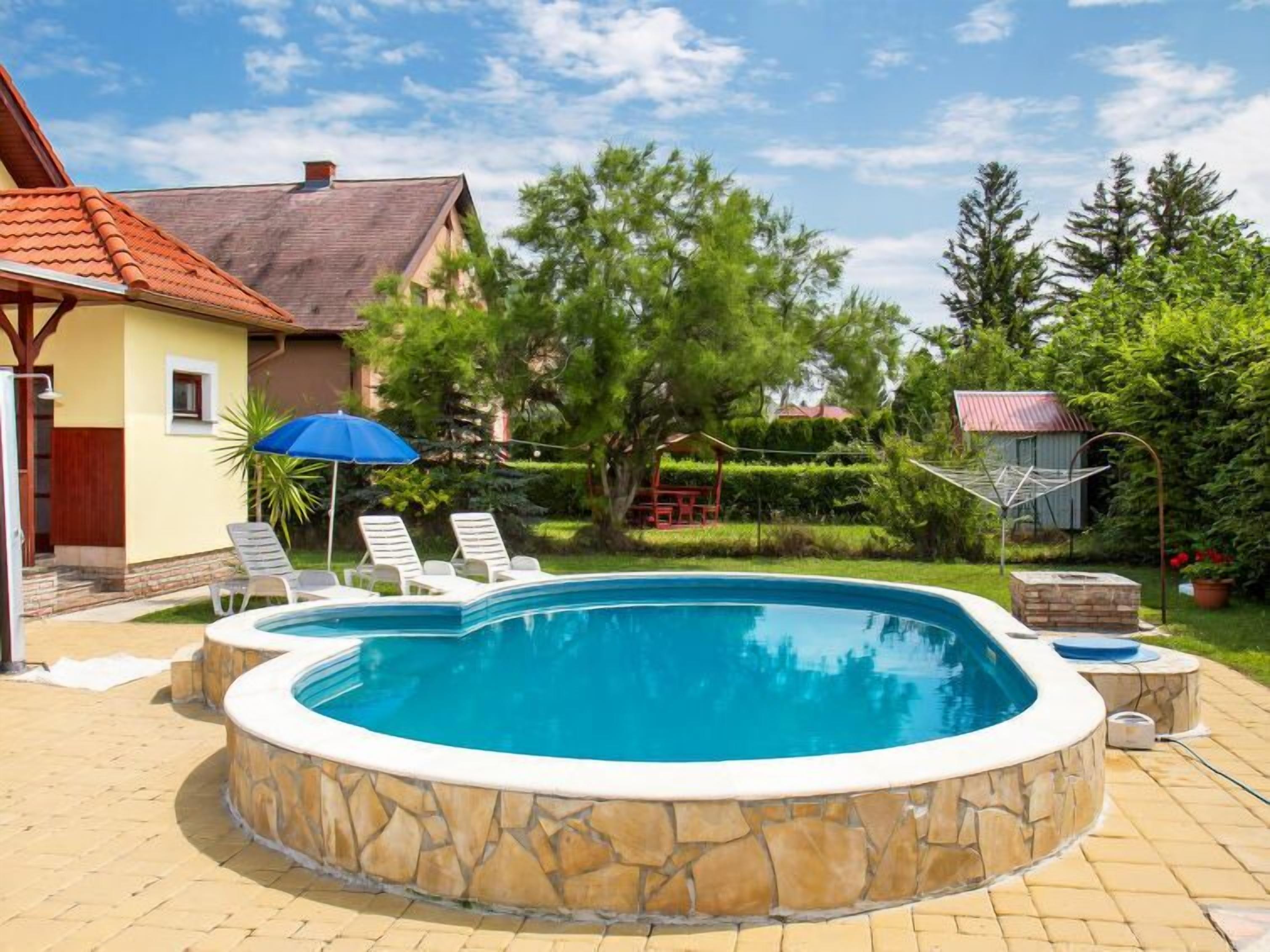 A swimming pool with a stone border, surrounded by a patio area with lounge chairs and a blue umbrella.