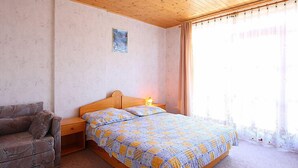 A bedroom with a wooden ceiling, a bed with a patterned bedspread, a nightstand, a wooden bench, and a window with curtains. - Yellow 1 by Interhome (Balatonmariafurdo)