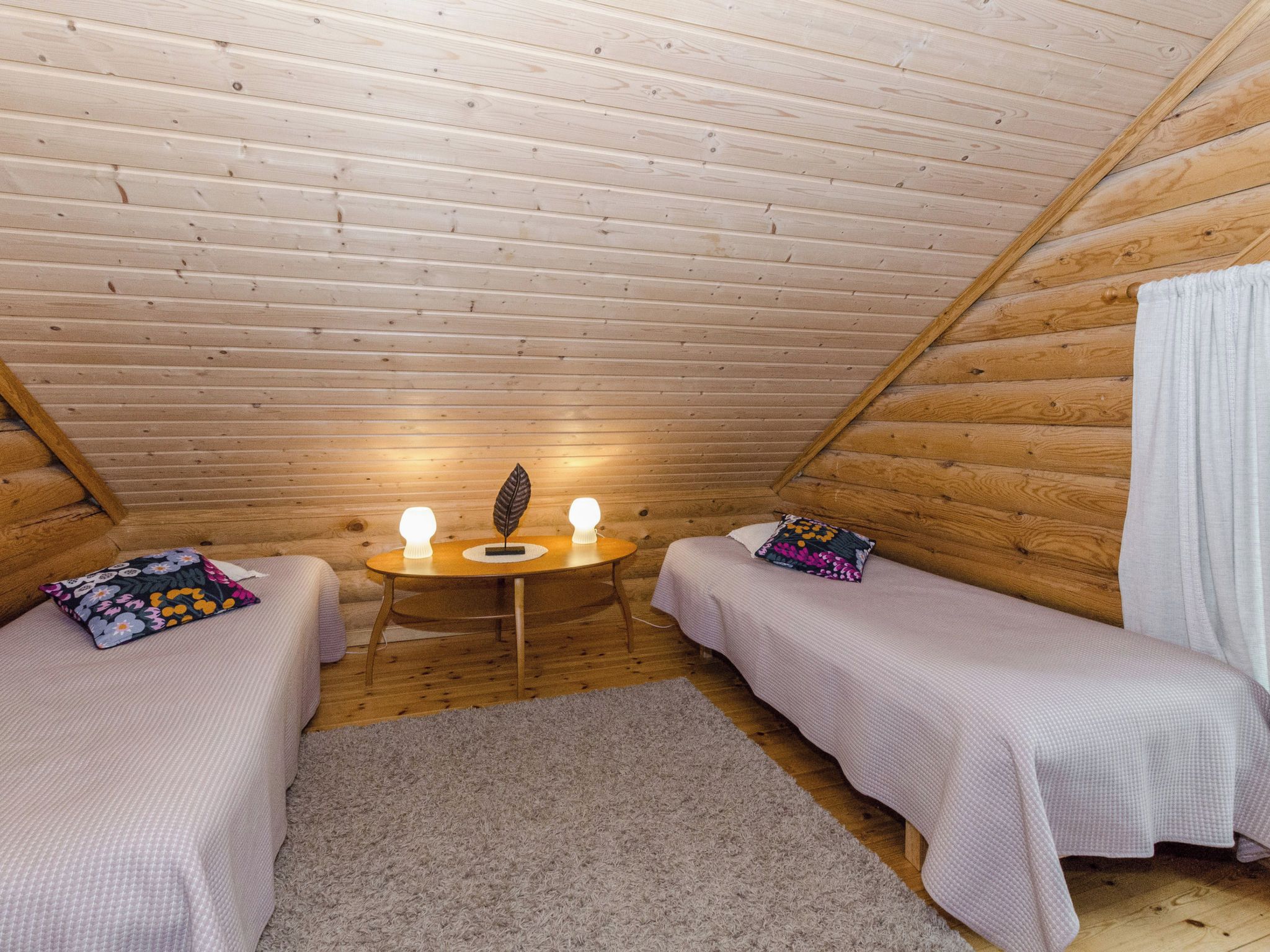 A small, cozy attic bedroom with two single beds, a small table with two lamps, and a wooden ceiling.