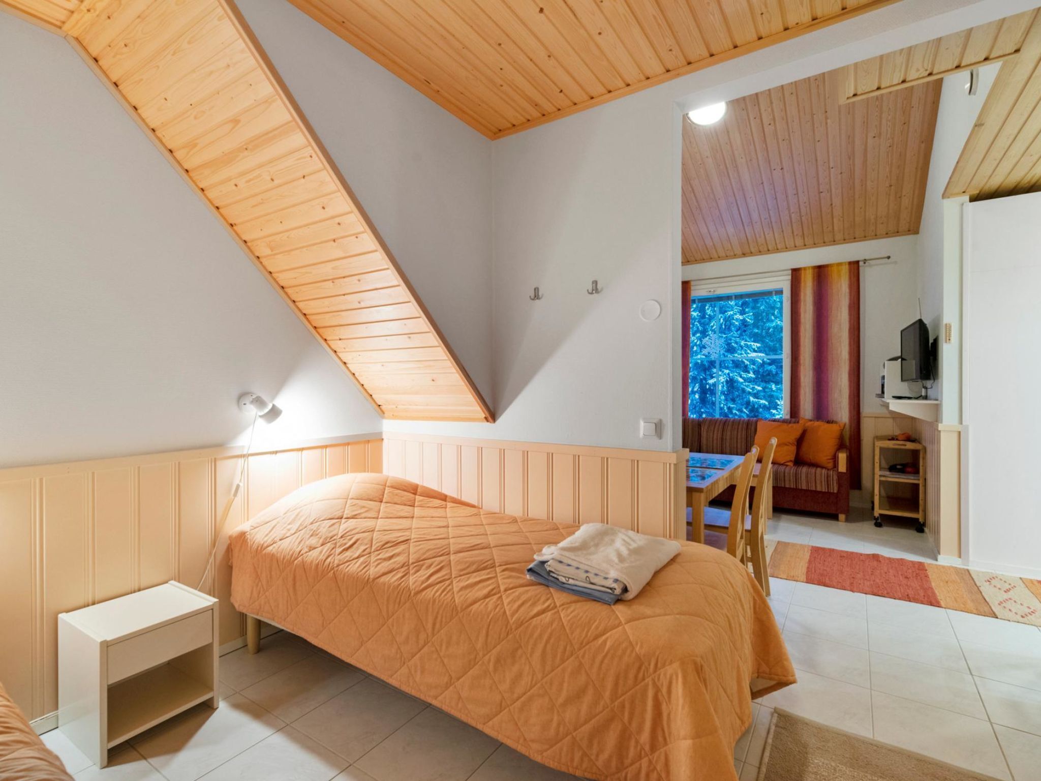 Une chambre avec un plafond en pente, un lit, une table de chevet, un bureau et une fenêtre donnant sur les arbres.