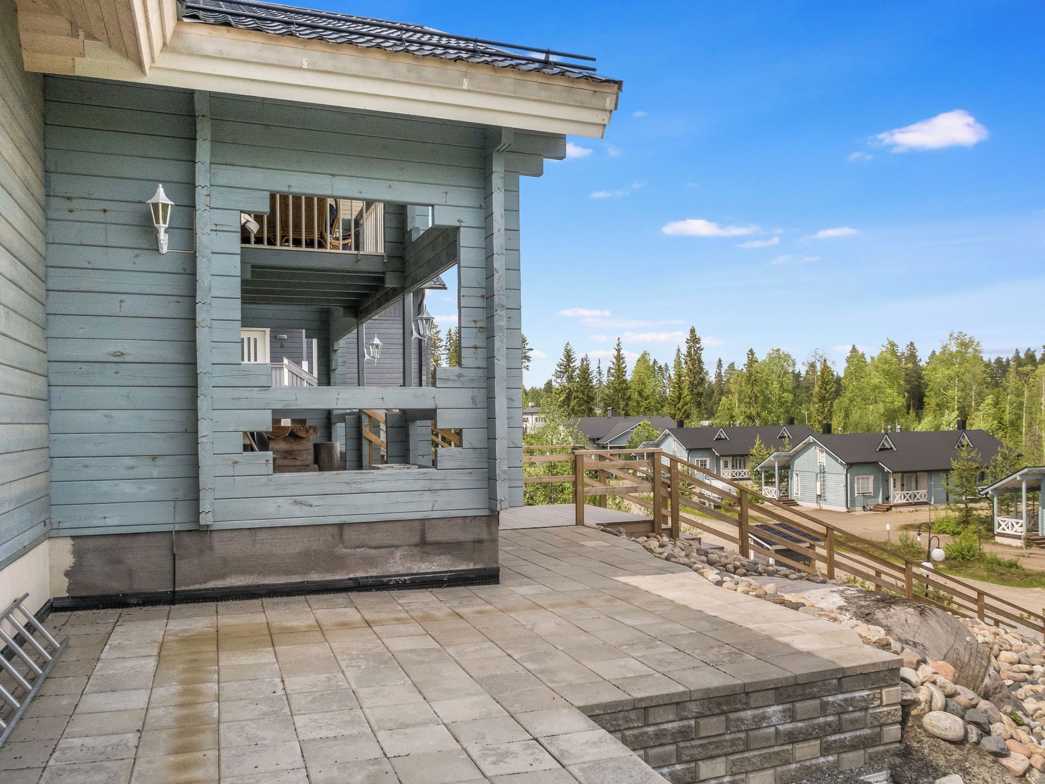 A two-story building with a balcony, a stone-paved patio, and a view of other houses in a forested area.