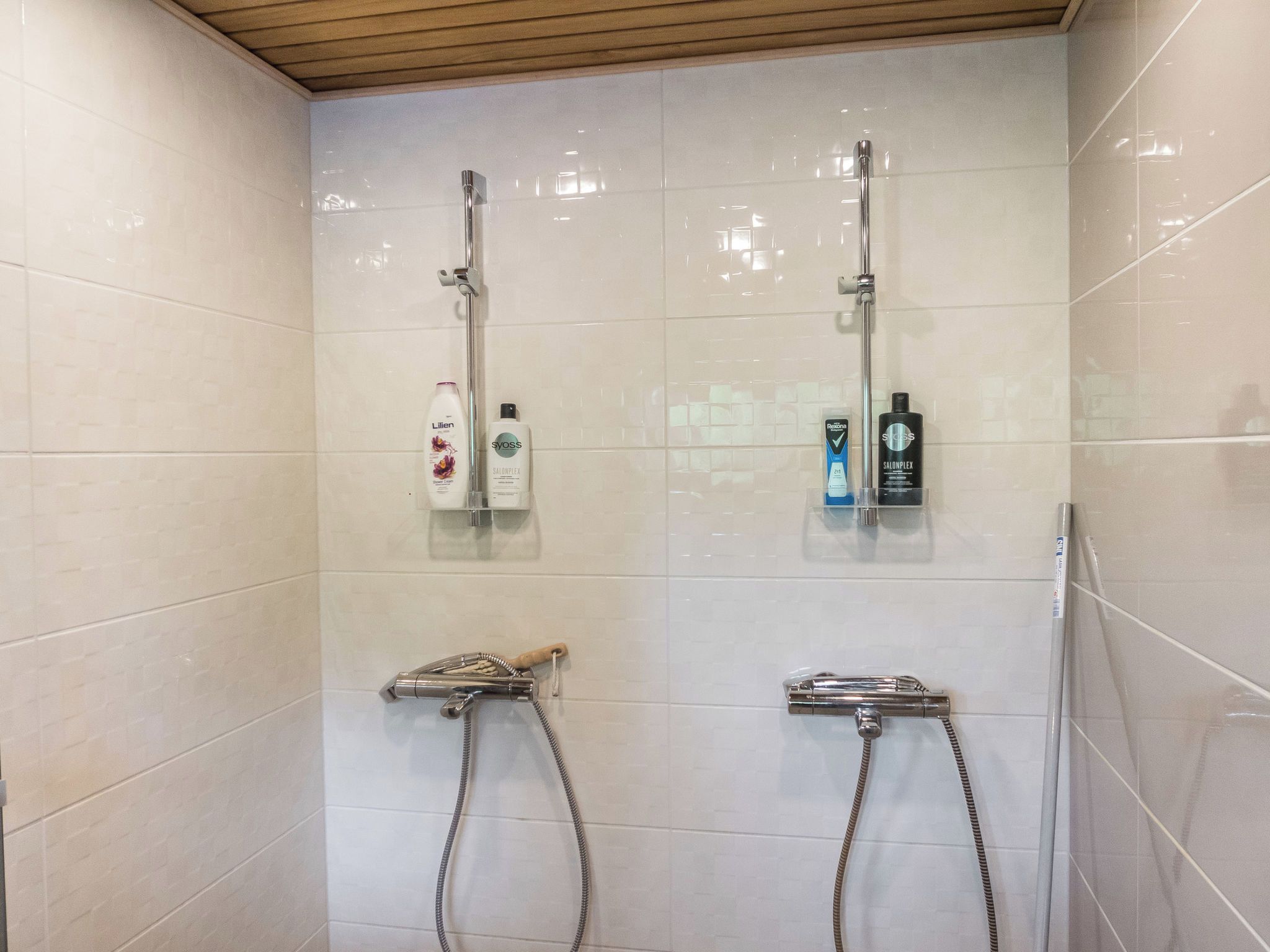 A shower area with two showerheads, a glass shelf with toiletries, and a wooden ceiling.
