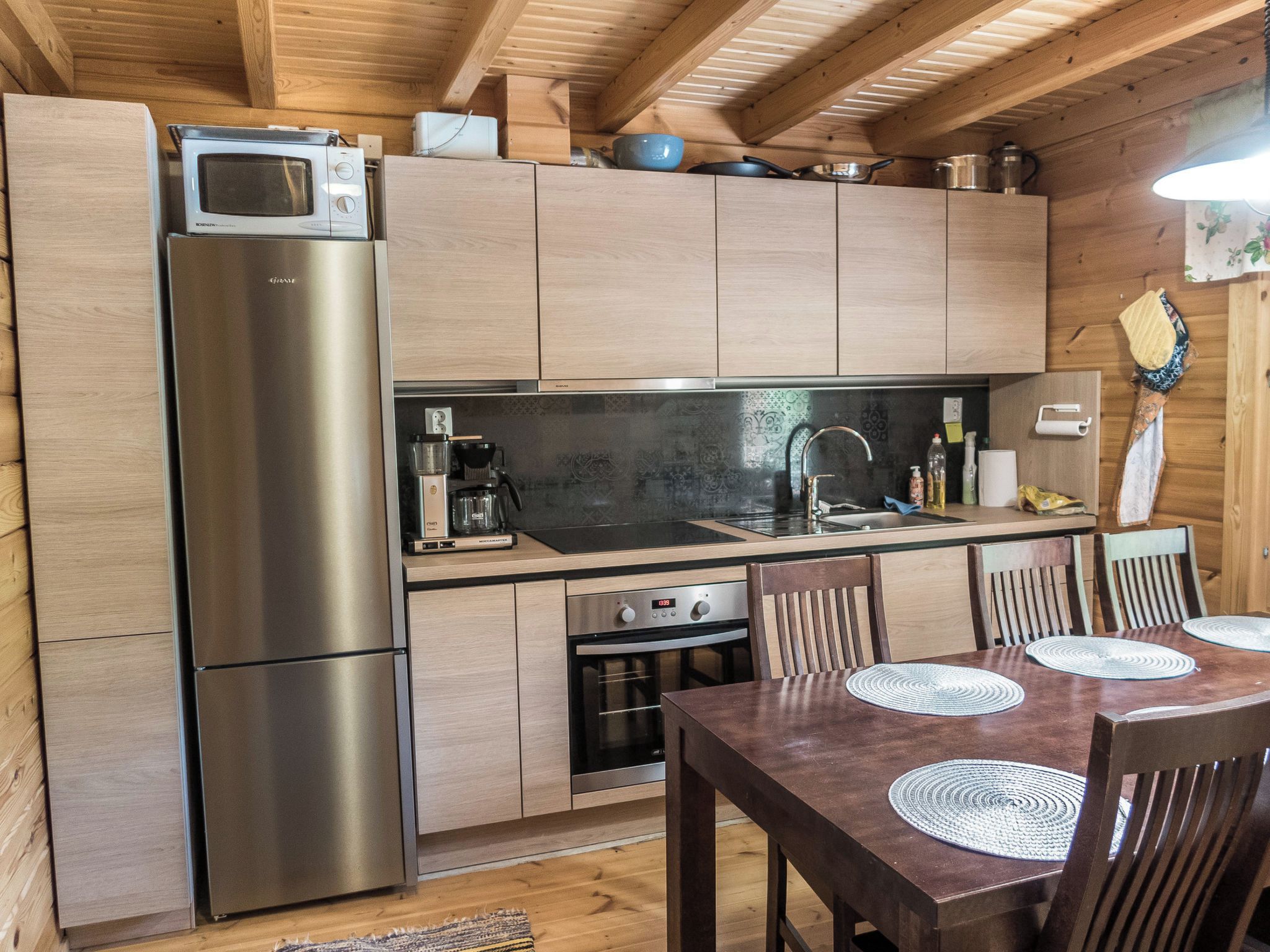 A compact kitchen with a stainless steel refrigerator, a microwave, and a coffee maker. There is a wooden dining table with chairs and a patterned placemat.