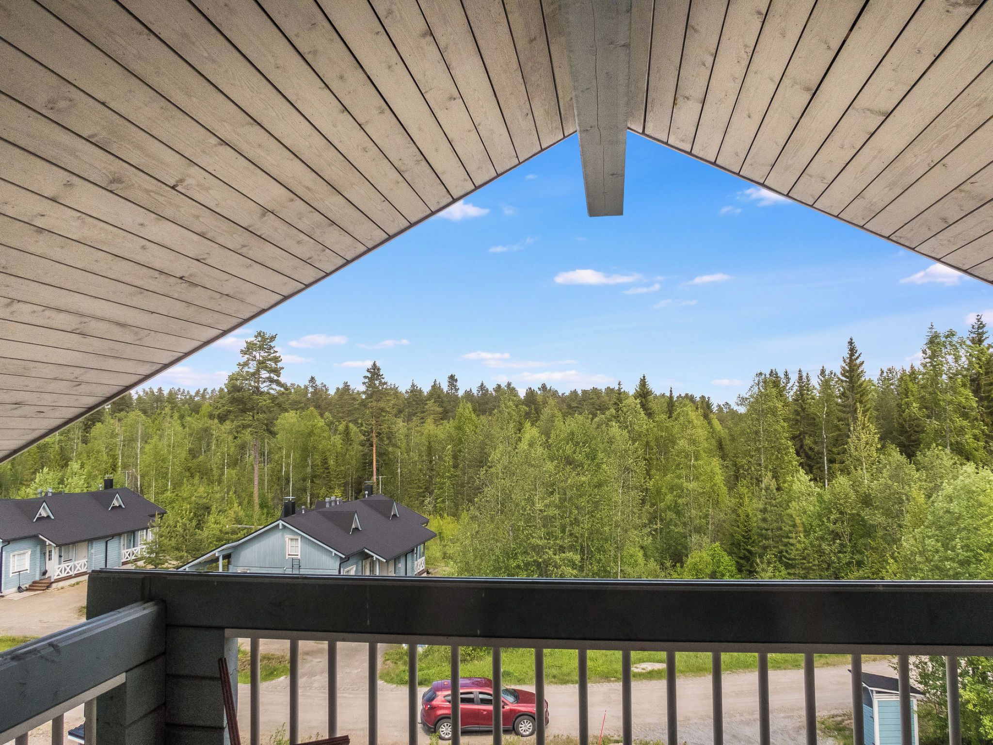 Näkymä parvekkeelta, jossa on puinen kaide. Parvekkeelta avautuu näkymä metsäiselle alueelle ja näkyy punainen auto, joka on pysäköitynä ajotielle.