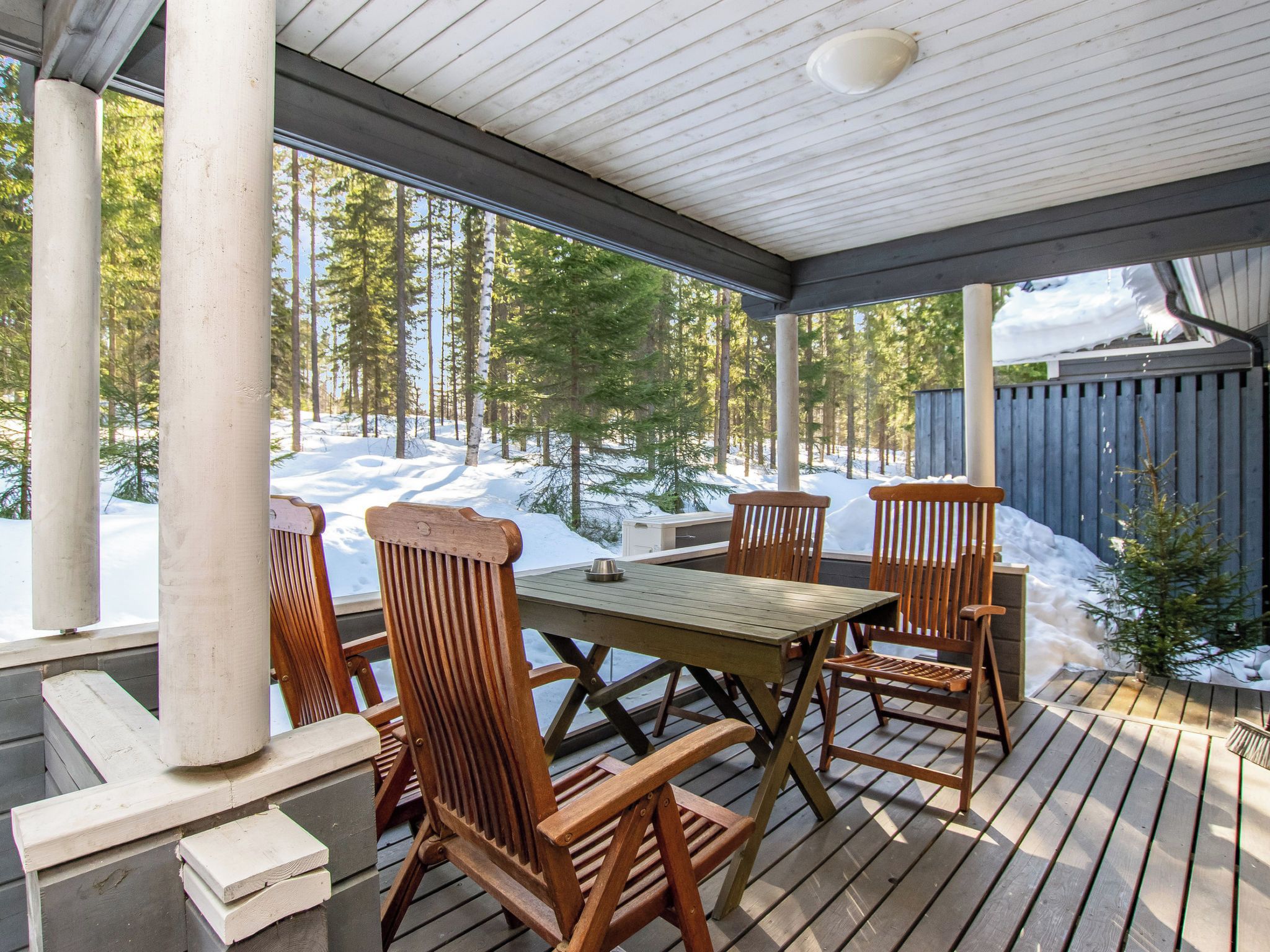 En träveranda med ett bord och stolar, med utsikt över ett snötäckt landskap med träd.