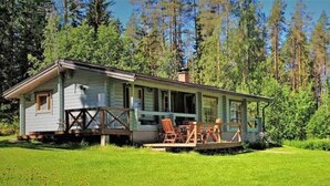 Pieni, yhden kerroksen mökki, jossa on kuisti ja ulkona oleva oleskelualue. Mökin ympärillä on nurmikko ja tiheä metsä.