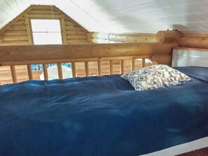 A wooden bed with a blue mattress and a patterned pillow in a cabin with a sloped ceiling and a window.