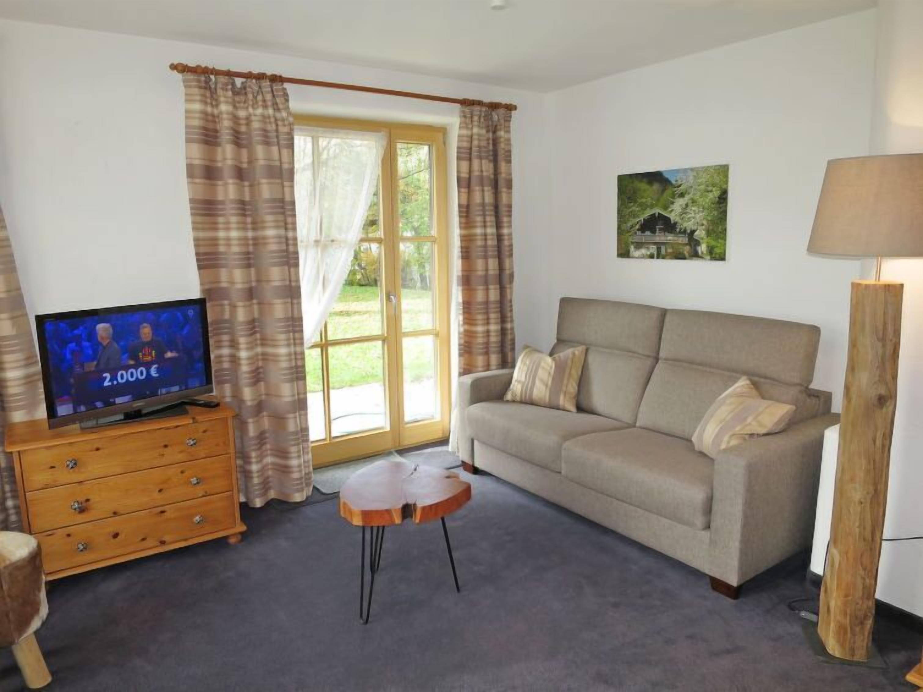 Ein Wohnzimmer mit einer Couch, einem Fernseher, einem Holzkasten, einem kleinen runden Tisch und einer Lampe.