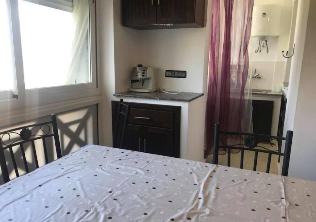 A compact room with a dining table, a kitchenette with a coffee maker, and a window with curtains.