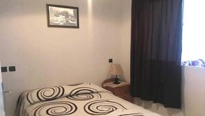 A bedroom with a bed, a nightstand, a lamp, a framed picture on the wall, and a window with curtains. - Residence Cite Jardin 4089 (Martil)