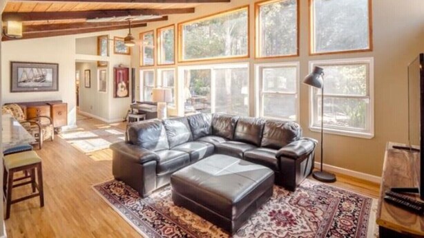 A spacious living room with a large sectional sofa, wooden flooring, and a high ceiling with exposed beams. - Pot Belly Beach House (Aptos)