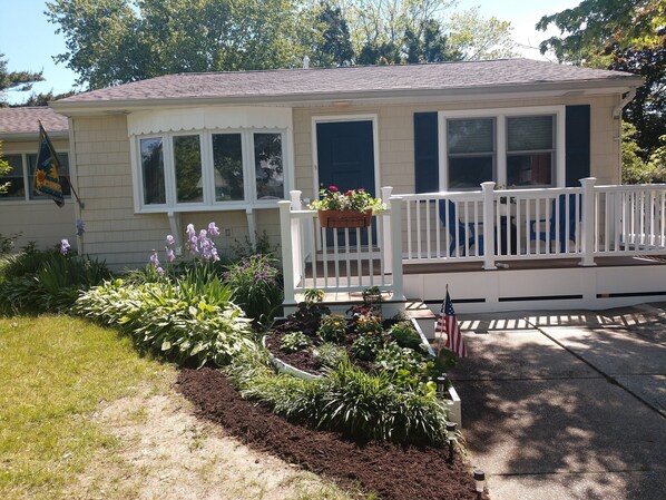 A single-story house with a front porch, a welcome sign, and a small garden with various plants and flowers.