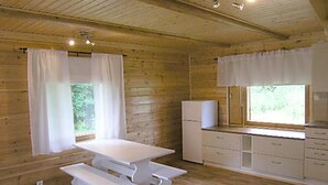 Interior de una cabaña de madera con una mesa de comedor, bancos, un refrigerador y una ventana con cortinas.