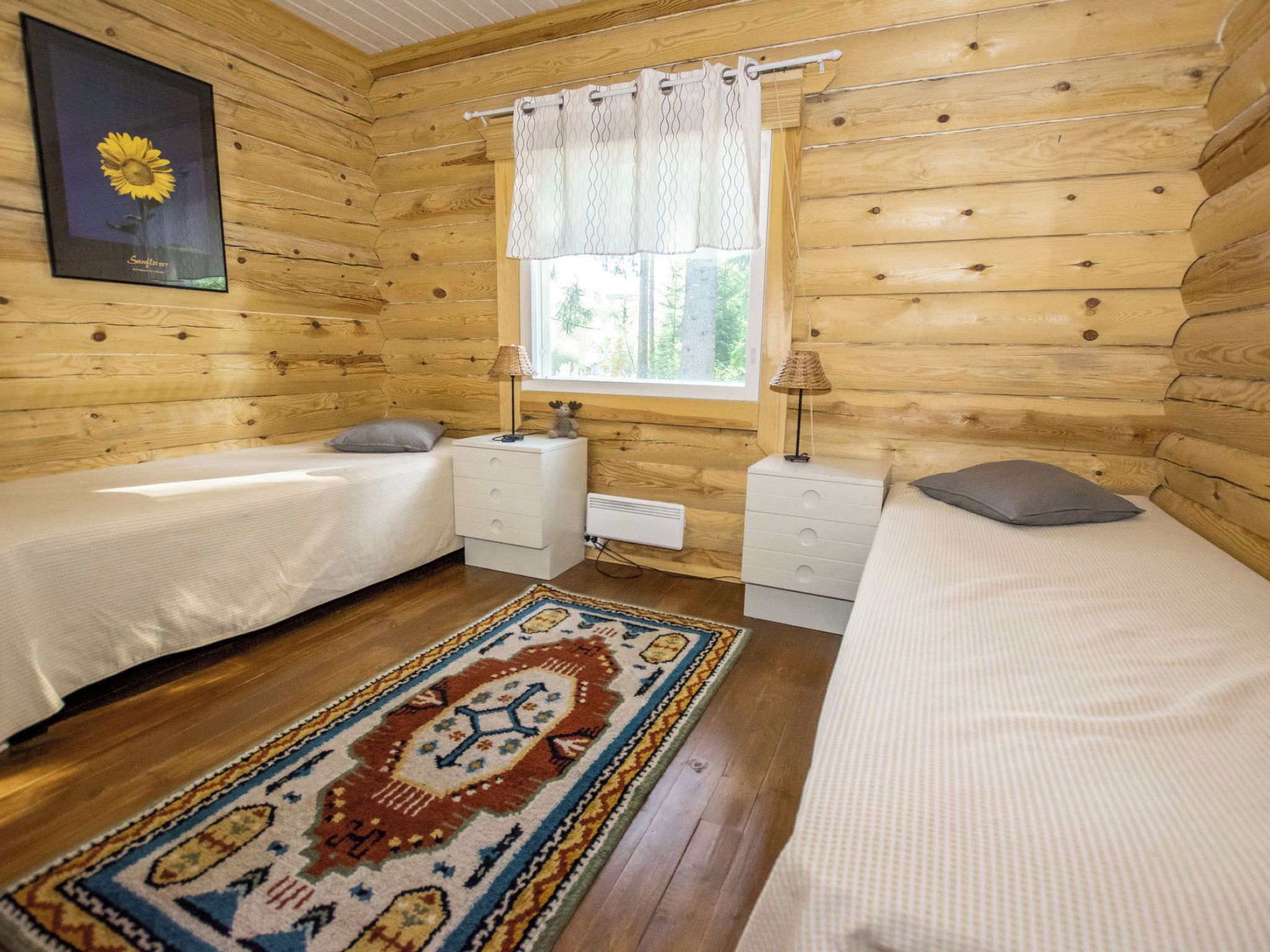 A wooden cabin bedroom with two beds, a window with curtains, a radiator, and a patterned rug.