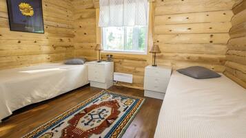 A wooden cabin bedroom with two beds, a window with curtains, a radiator, and a patterned rug.
