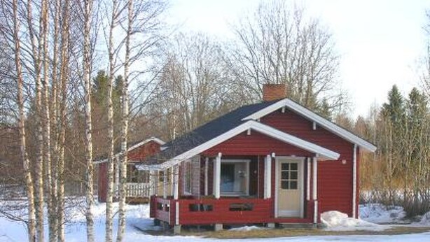 Pieni punainen mökki, jossa on kuisti, ja sen ympärillä on lunta ja puita.