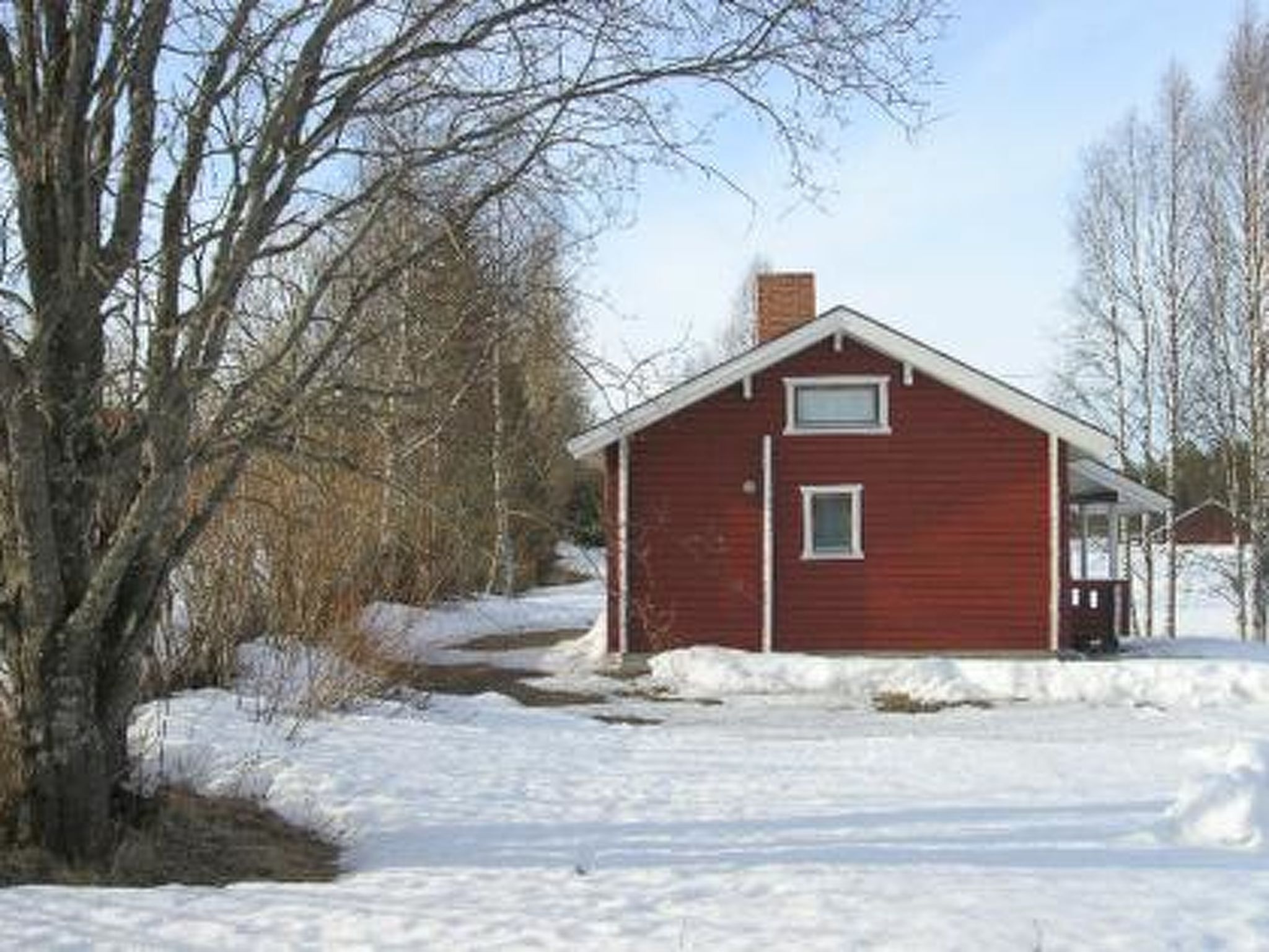 Ein rotes Holzhaus mit Satteldach und Schornstein, umgeben von verschneitem Boden und kahl stehenden Bäumen.