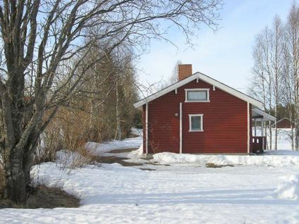 A red wooden house with a gabled roof and a chimney, surrounded by snow-covered ground and leafless trees.