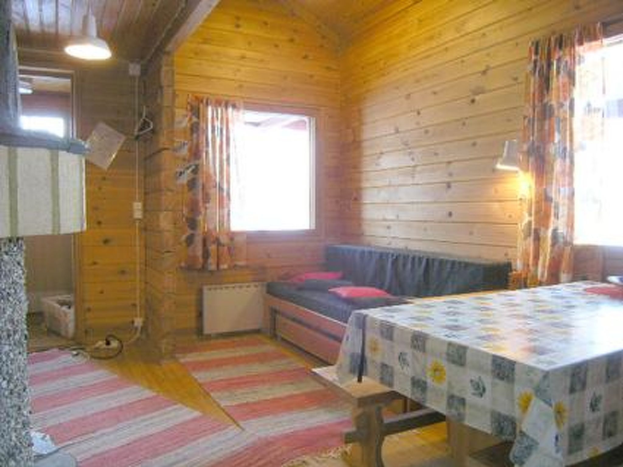 A cabin with a wooden interior, a bed with a red pillow, a table with a floral tablecloth, and a window with curtains.