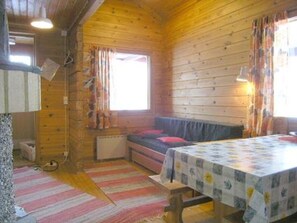 A cabin with a wooden interior, a bed with a red pillow, a table with a floral tablecloth, and a window with curtains.