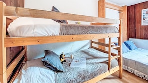 Ein Etagenbett mit unterem und oberem Schlafplatz, beide mit Kissen und Decke. Auf dem unteren Bett befindet sich ein kleiner Tisch mit einem Teddybären und einem Buch.