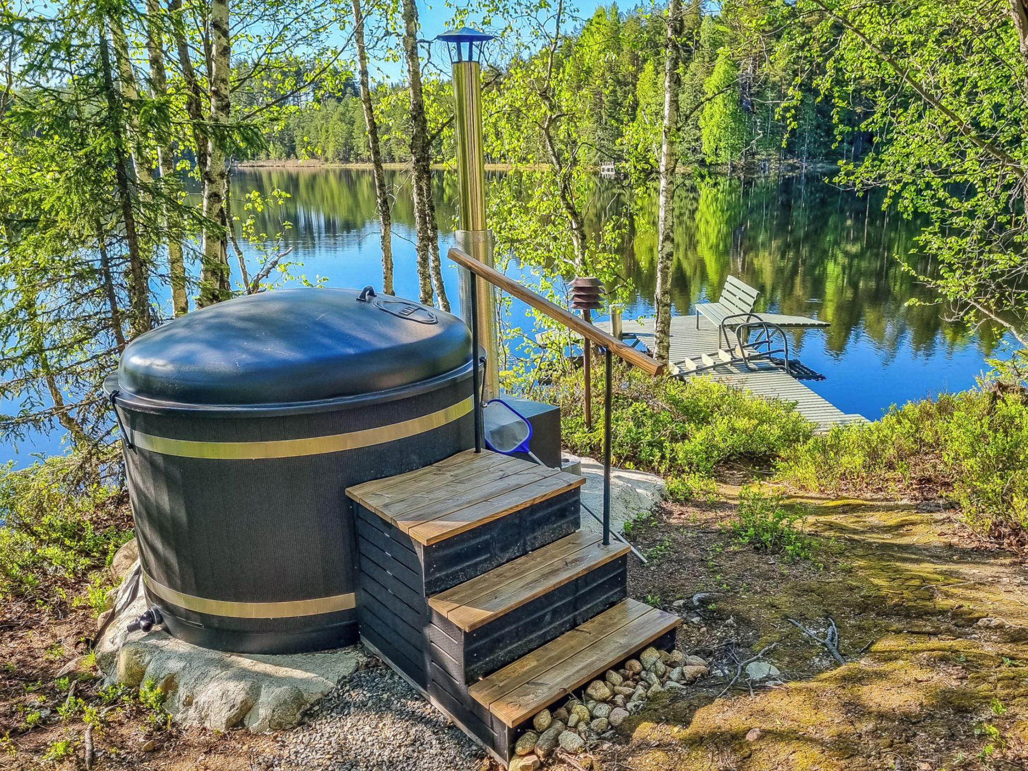 Een sauna met een houten terras en een trap, gelegen in een bebost gebied aan een meer.