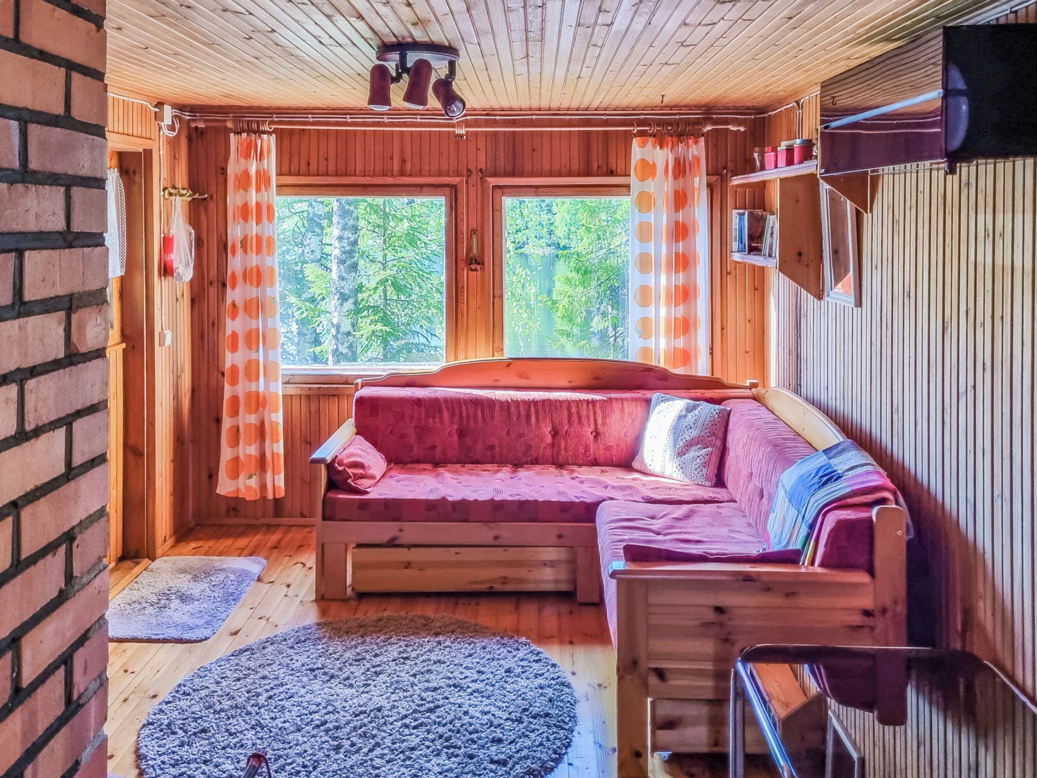 A cozy cabin interior with a wooden floor, a red sofa, a small rug, and a window with curtains.