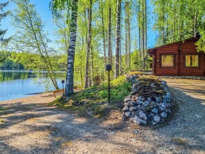Une cabane au bord d’un lac, entourée de bouleaux, avec un mur de pierres et un chemin de gravier.