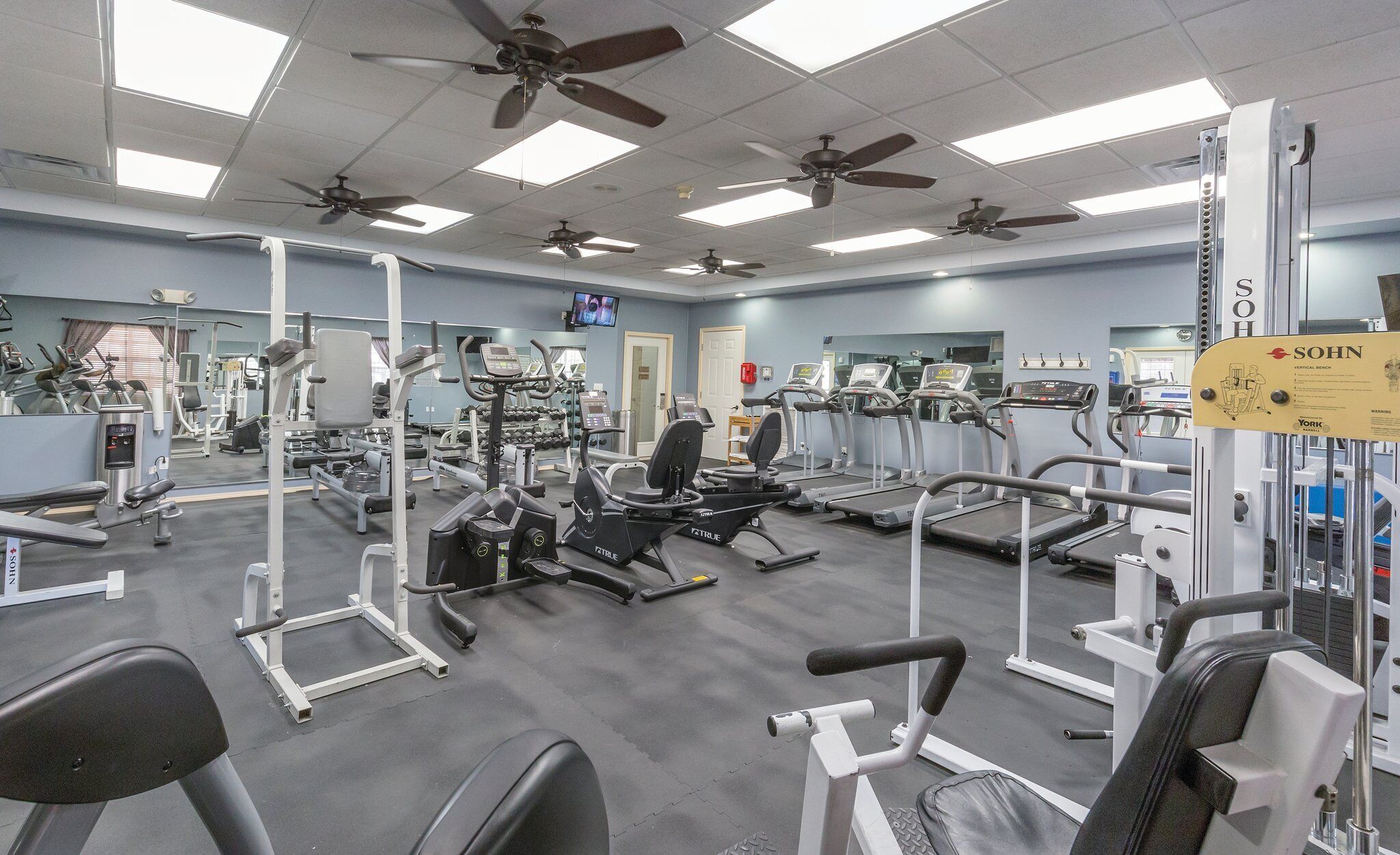 A gym with various exercise machines, ceiling fans, and a sign for a vertical bench.