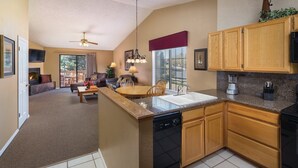 A modern kitchen with wooden cabinets, a dishwasher, and a granite countertop. There is a dining area with a table and chairs, and a living room with a fireplace and a ceiling fan.