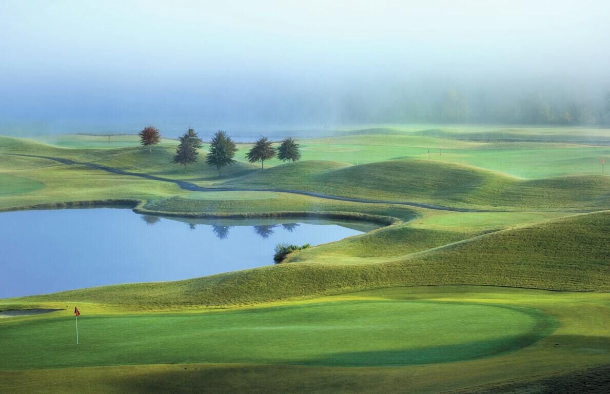 A golf course with rolling green hills, a pond, and a flag.