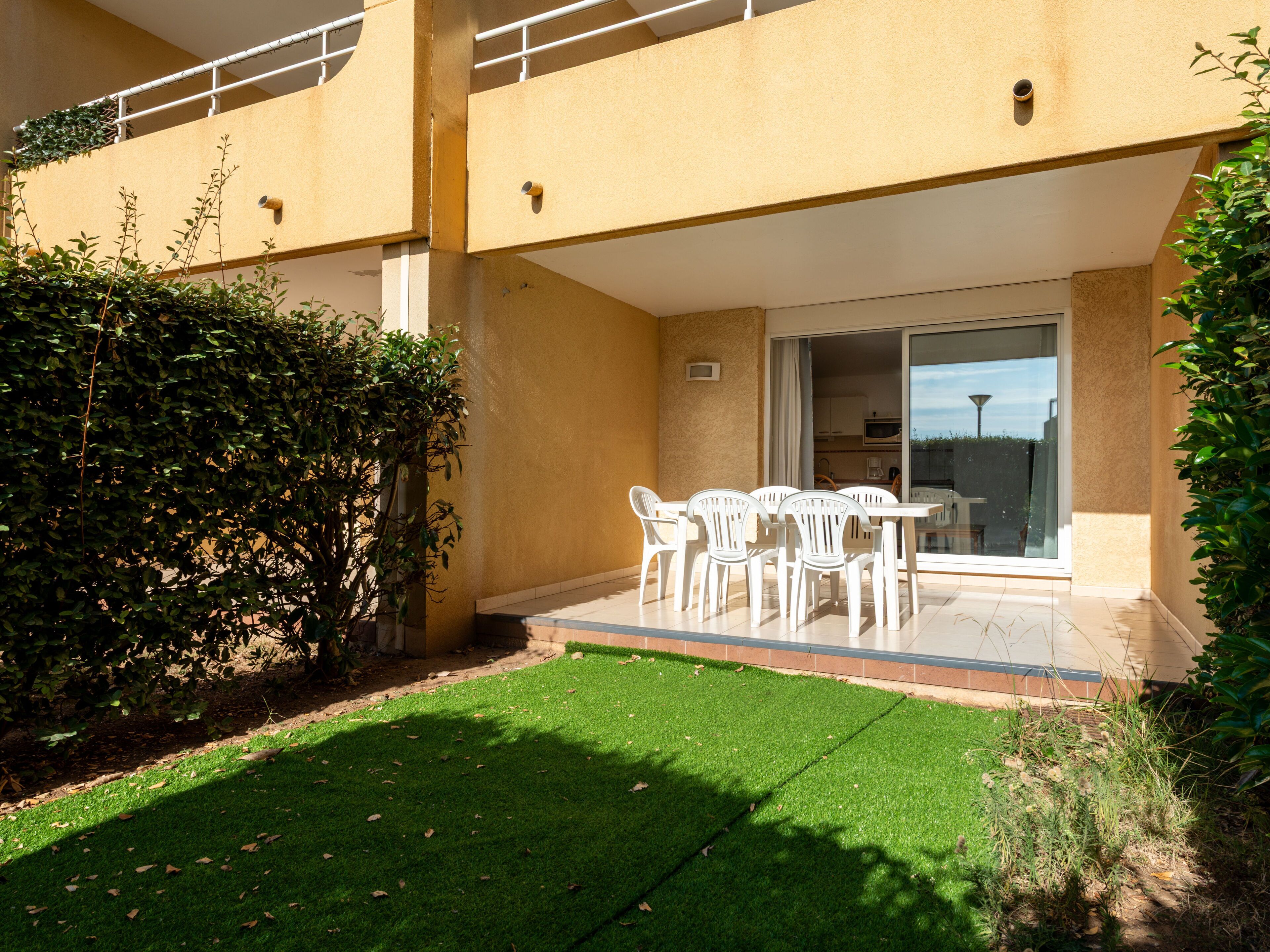 Une terrasse avec une table et des chaises, une porte vitrée donnant sur un balcon et une pelouse bien entretenue.