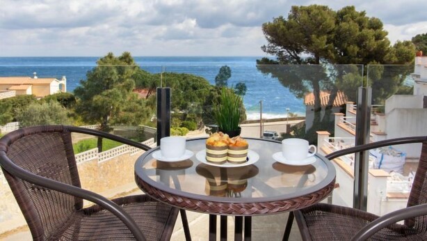 A balcony with wicker chairs, a glass table, and a view of the sea and trees.