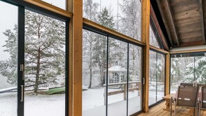Un gran ventanal da a una terraza de madera con vistas a un paisaje nevado y una cabaña.