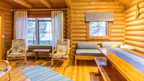 Interior de una cabaña de madera con un sofá, dos sillones, una mesa y un ventanal con cortinas.