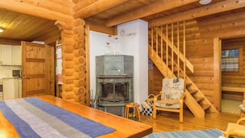 A log cabin interior with a wooden dining table, a fireplace, a staircase, and a kitchen area.