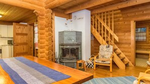 Interior de una cabaña de troncos con una mesa de comedor de madera, una chimenea, una silla y una escalera.