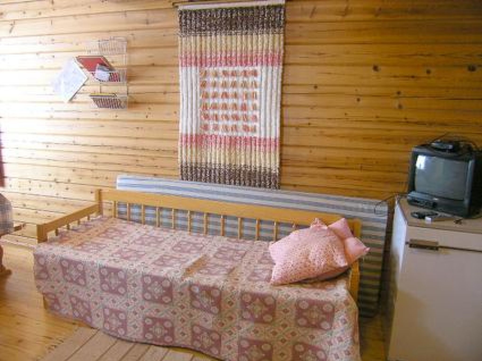 Een knus, klein kamertje met een houten vloer, een bed met een beddeken met een patroon, een televisie op een dressoir, en een gordijn dat aan de muur hangt.