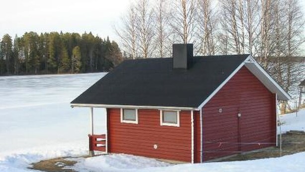 A small, red wooden cabin with a black roof, a chimney, and a small porch with a bench.