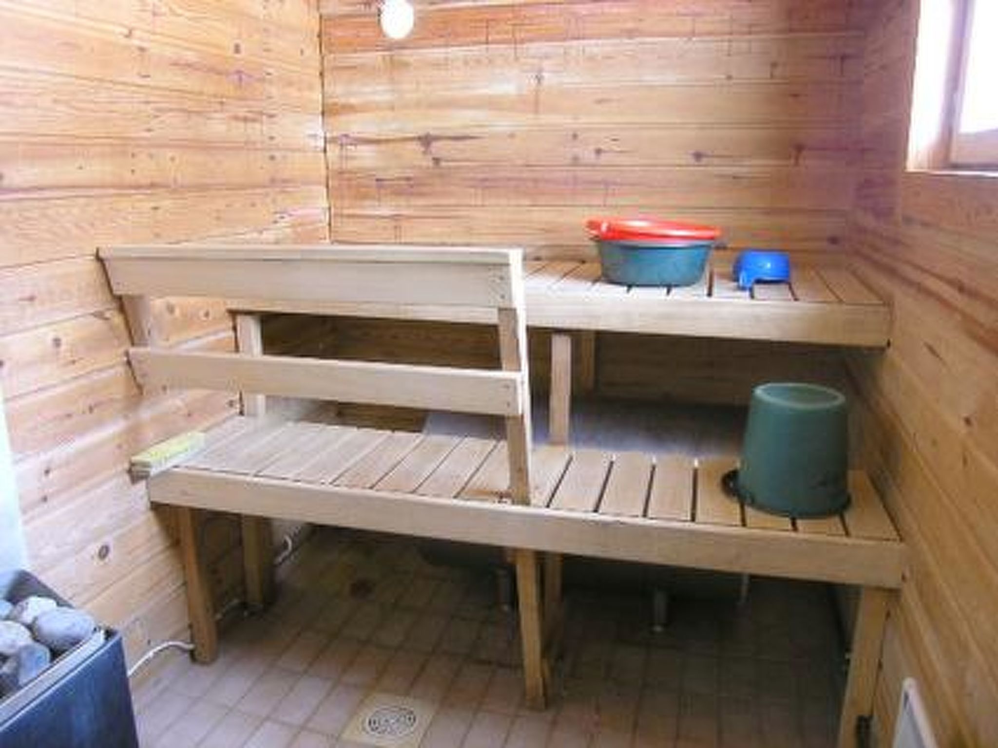 A small wooden cabin with a bench, a blue bucket, and a red container on a shelf.